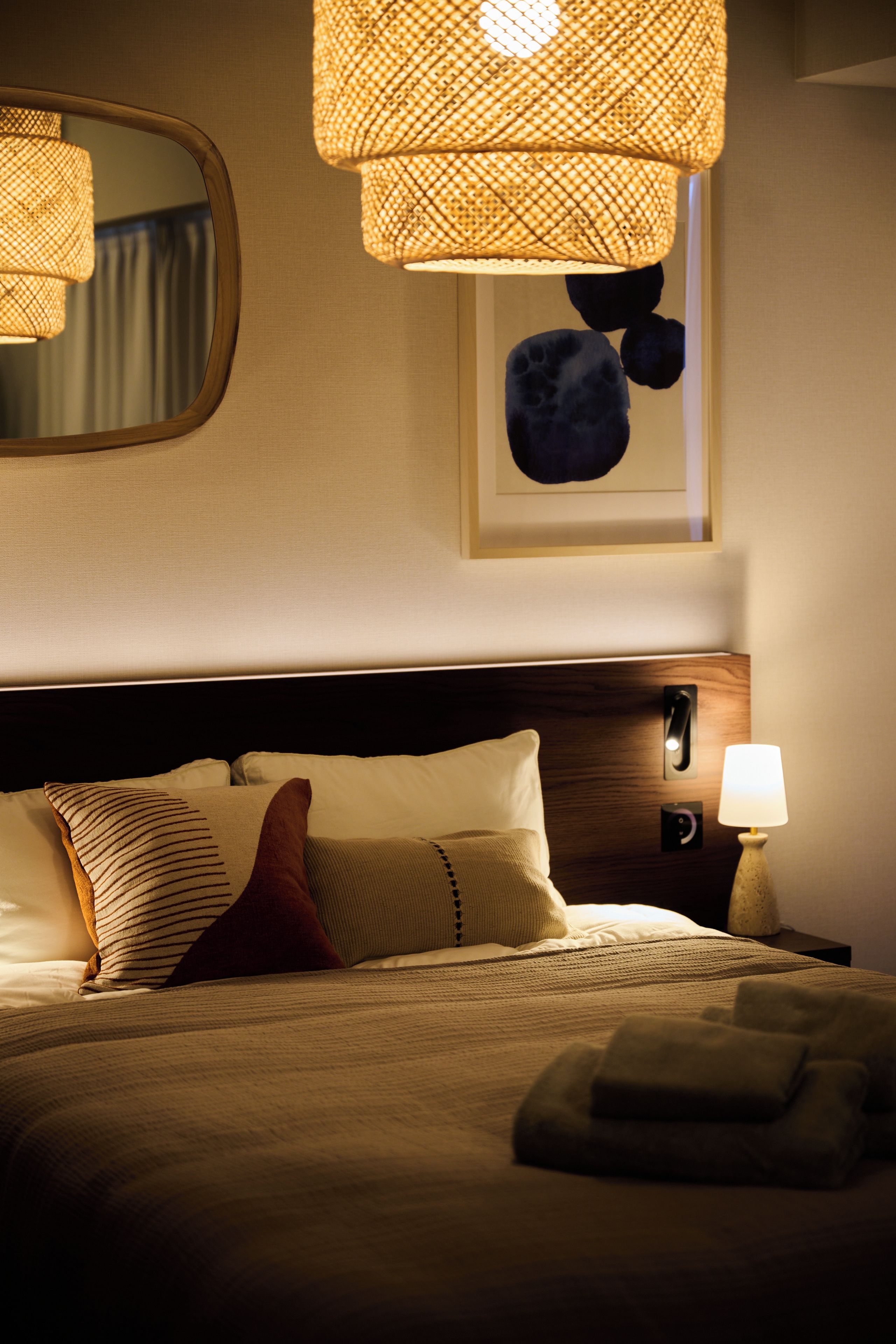 Een hotelkamer met een bed, kussens, een bedlampje, een spiegel aan de muur en een kunstwerk in een lijst.
