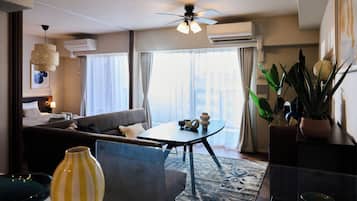 A modern hotel room with a sofa, a dining table, a ceiling fan, and a large window with curtains.