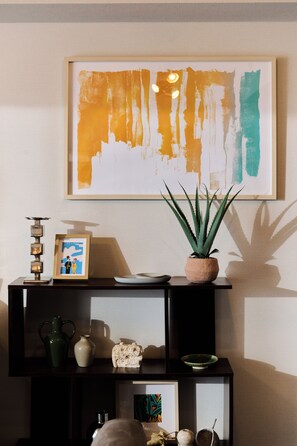 Una habitación con un cuadro abstracto colgado en la pared, una planta en maceta y varios objetos decorativos en una estantería.