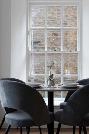 A dining area with a table set for two, two grey chairs, and a large window with a stone wall behind it. - Modern Apartment in City Centre (Edinburgh)