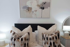 Un dormitorio con cama, almohadas, una mesita de noche, lámpara y un cuadro abstracto colgado en la pared.