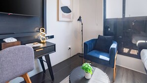 A modern living room with a desk, a blue armchair, a round coffee table with a plant, a floor lamp, and a large window. - Deluxe Twin Room - Sleeps 3 (Greenacre)