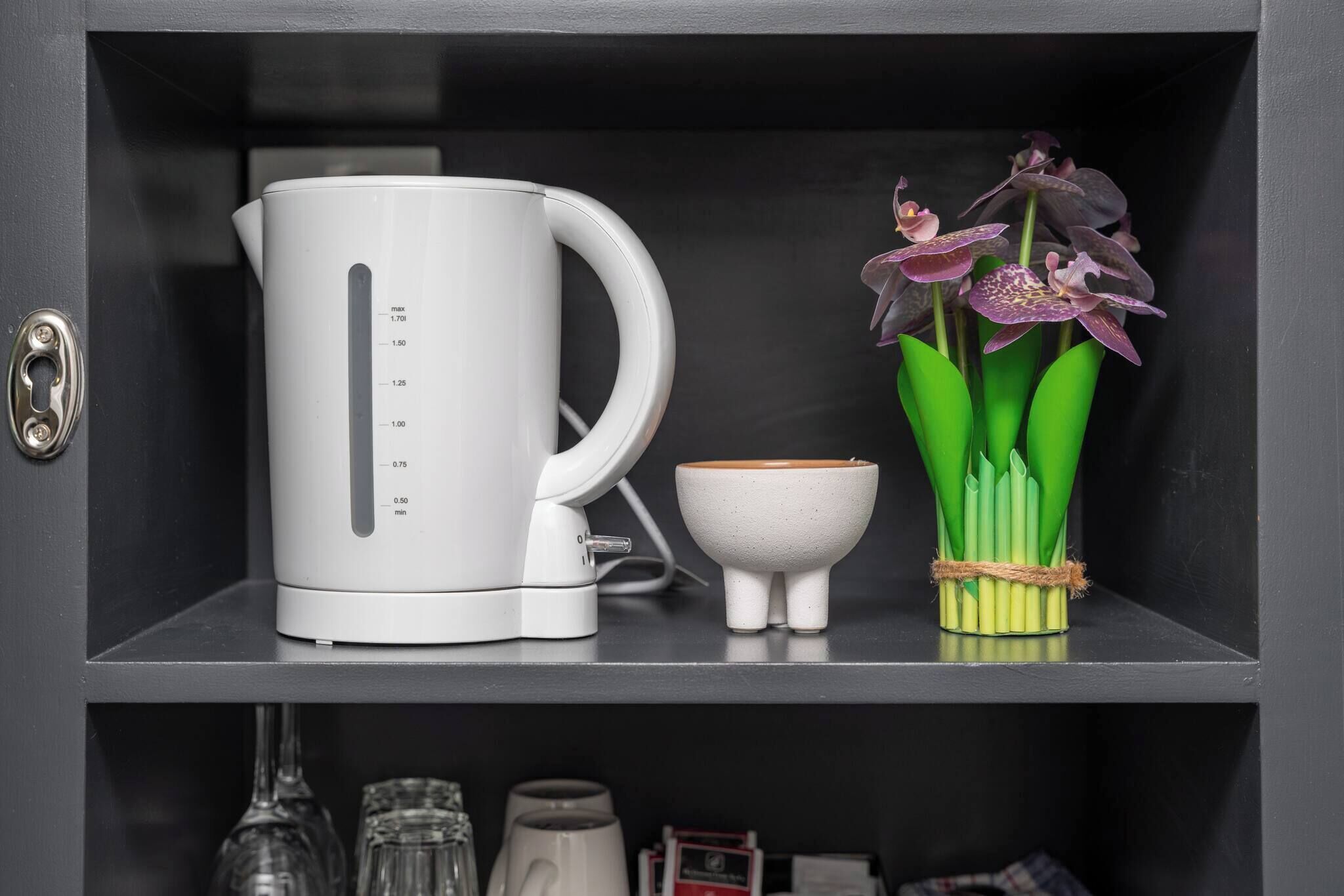 Eine weiße elektrische Kanne, eine kleine weiße Schale und ein Strauß violetter Orchideen auf einem schwarzen Regal.