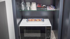 Un microondas con un plato de aperitivos encima, una cafetera y una estantería con cristalería y bocadillos.