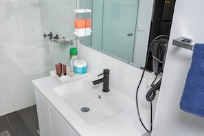 Un baño moderno con lavamanos blanco, espejo, secador de pelo y artículos de aseo.