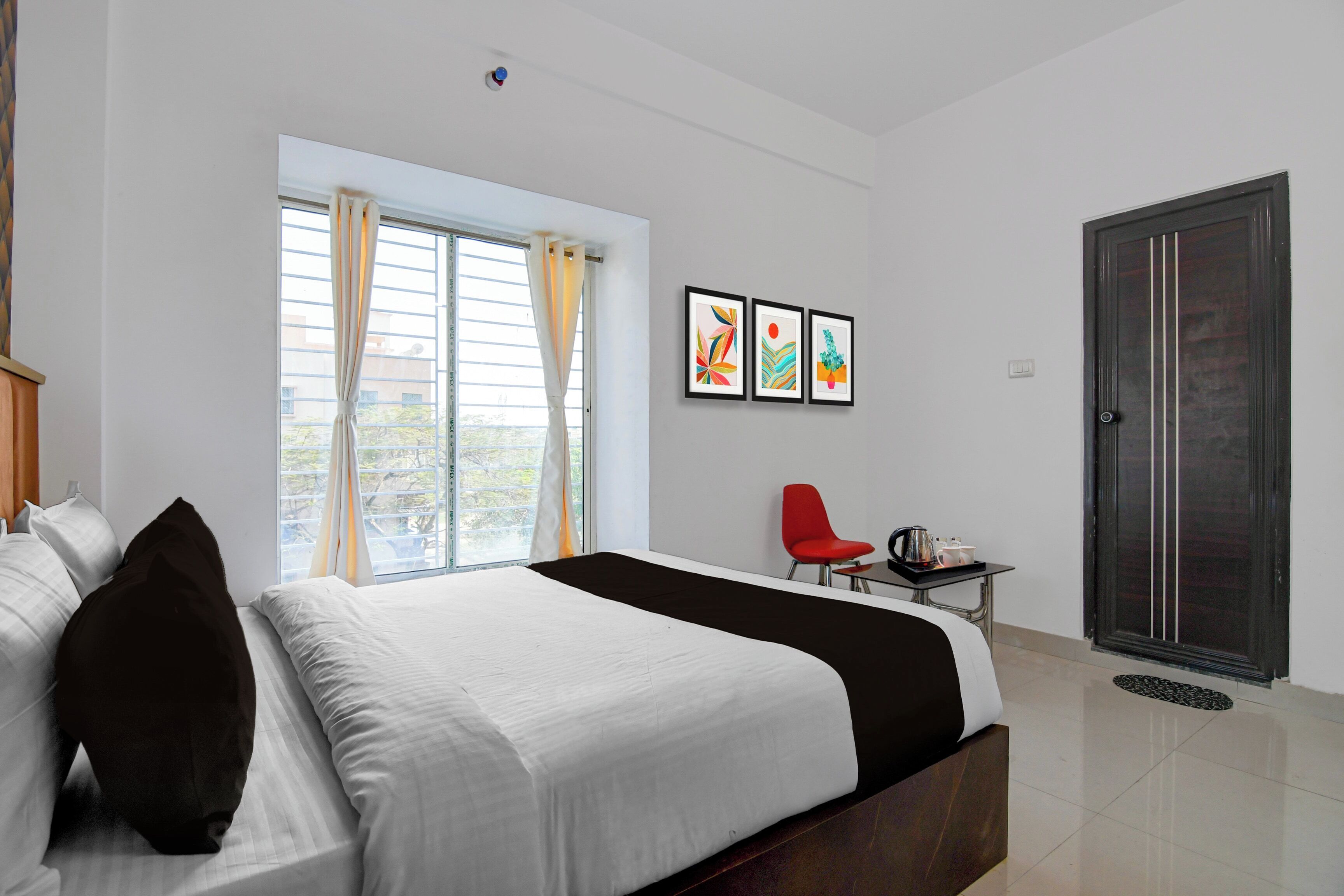 Une chambre d’hôtel avec un lit, une fenêtre avec des rideaux, une chaise rouge, une petite table avec un plateau, et une porte.
