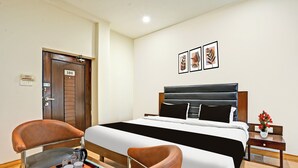 Habitación de hotel con cama, escritorio con bandeja, silla, mesita de noche con un florero y una puerta con placa numérica.