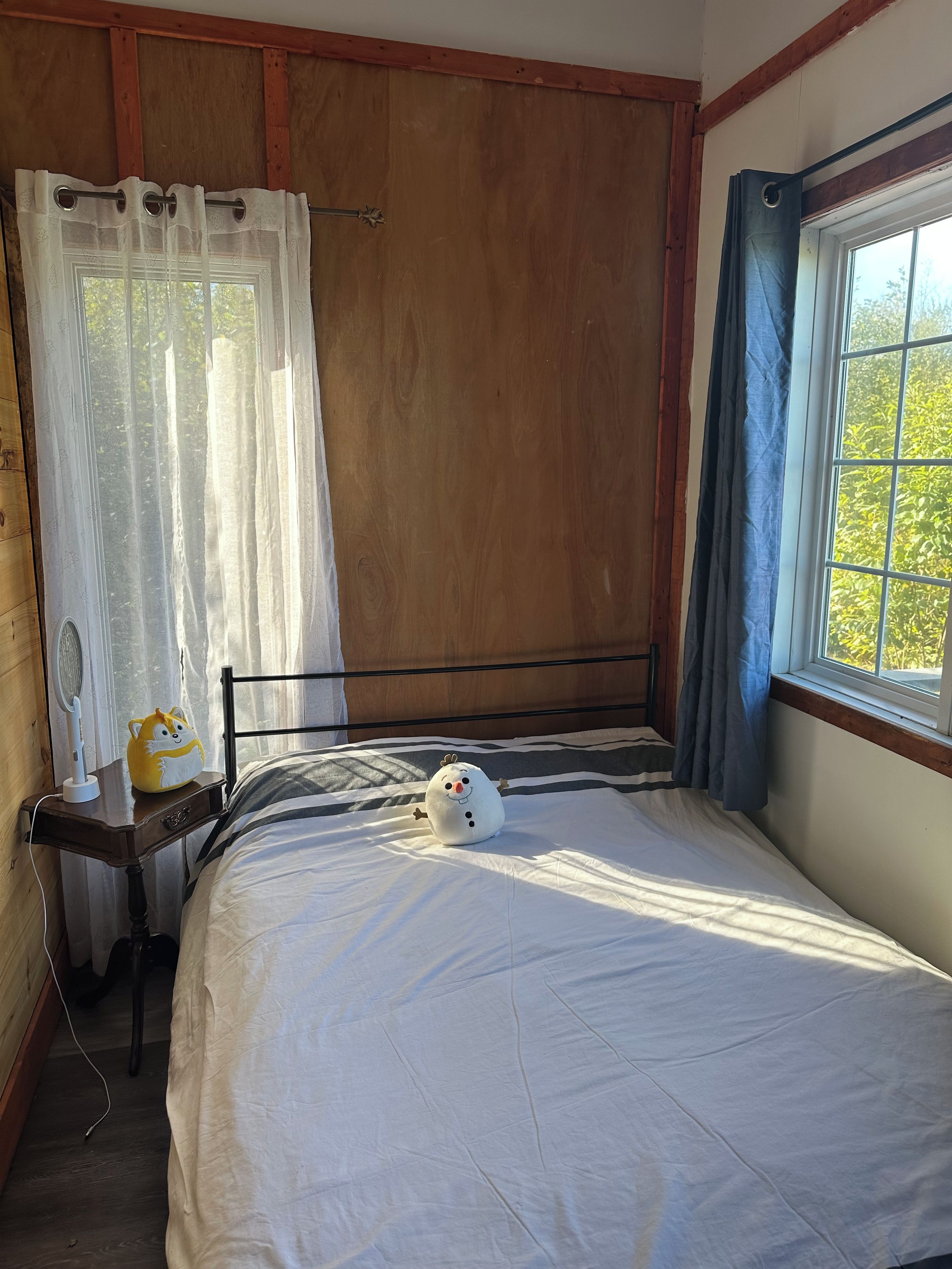 Une chambre à coucher avec un lit, une table de chevet avec une lampe, une fenêtre avec des rideaux et une vue sur la verdure à l’extérieur.