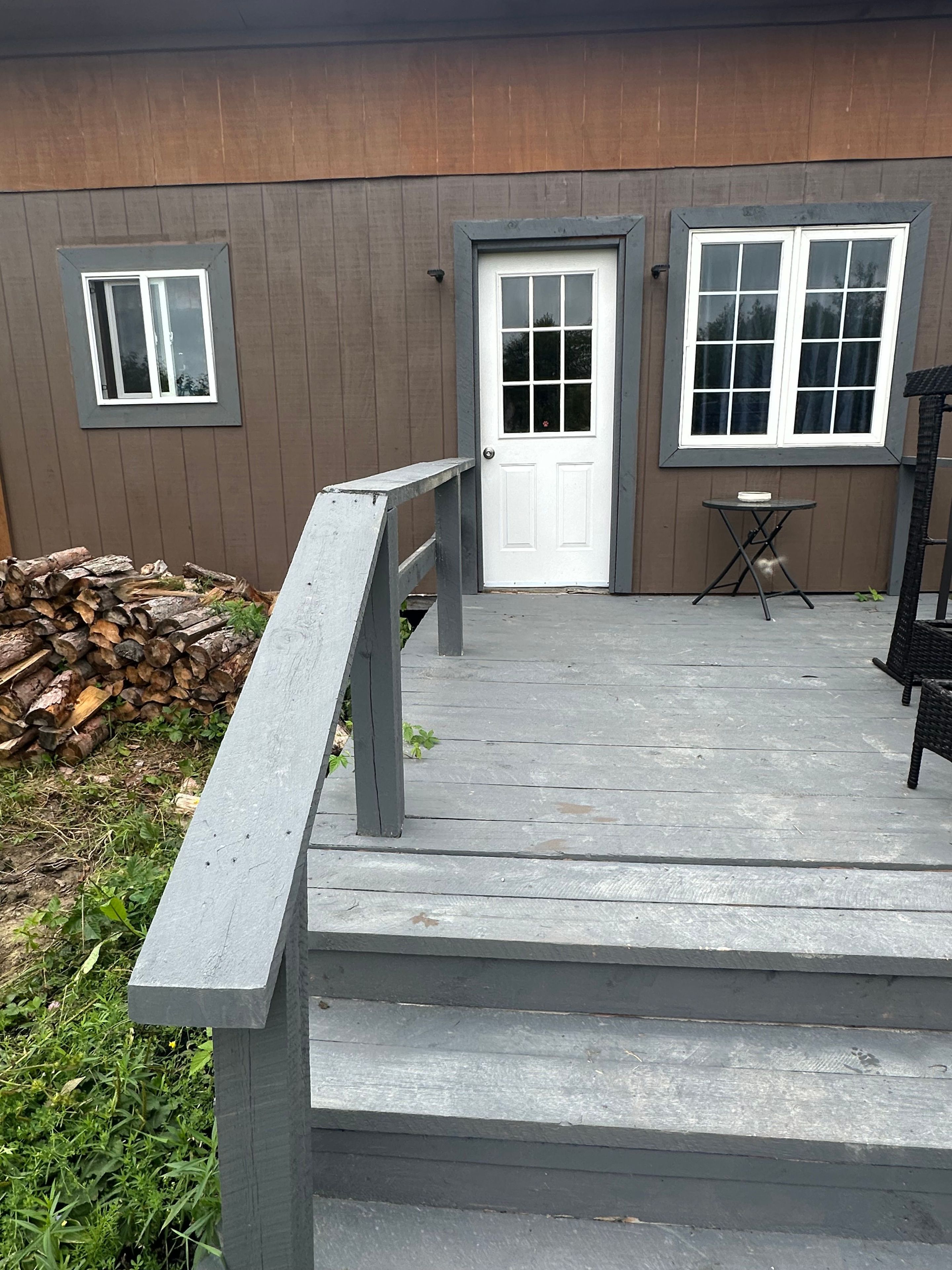 A wooden deck with a gray railing leading to a front door, a small table, and a stack of firewood.