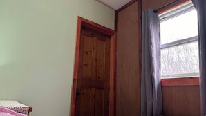 A bedroom with a wooden door, a window with curtains, and a bed with a pink and white quilt. - Fox Den French river (Noëlville)