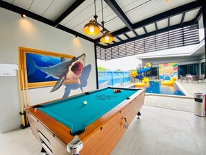 Une table de billard ornée d’une fresque représentant un requin, une piscine et un toboggan en arrière-plan.