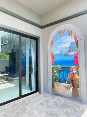 Ein Zimmer mit einer großen Glasdoor, die zu einem Außenbereich führt, einer Wandmalerei mit einer Küstenansicht und einem Balkon mit Geländer und Pflanzgefäßen.