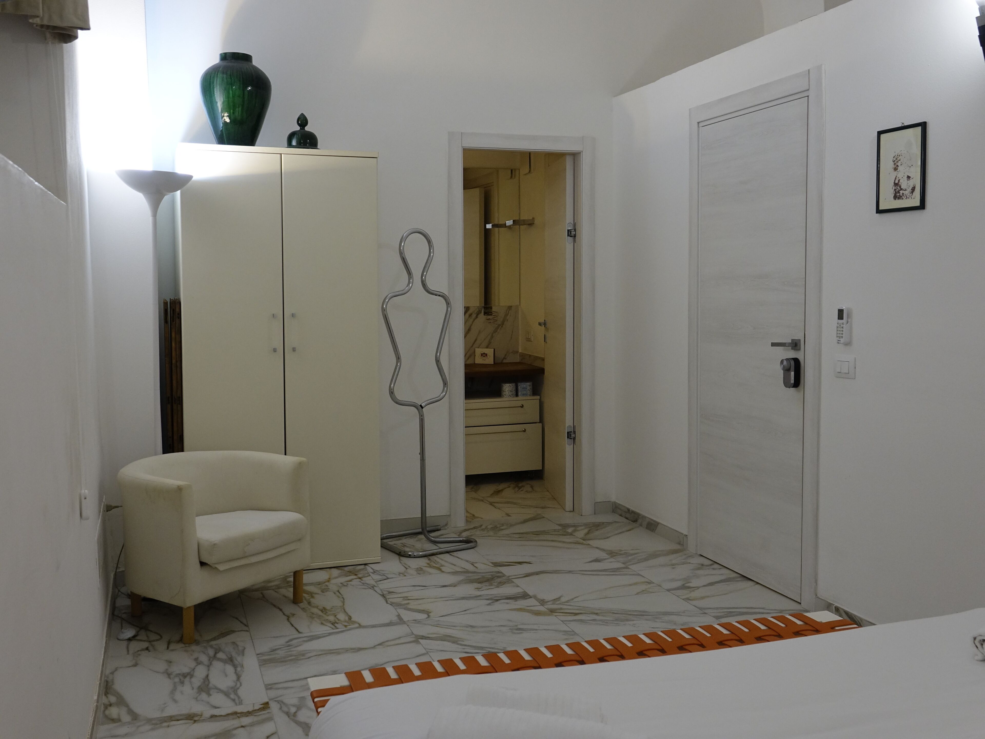 Ein Schlafzimmer mit Bett, Schrank, Stuhl, Stehlampe und einem durch eine offene Tür einsehbaren Badezimmer.