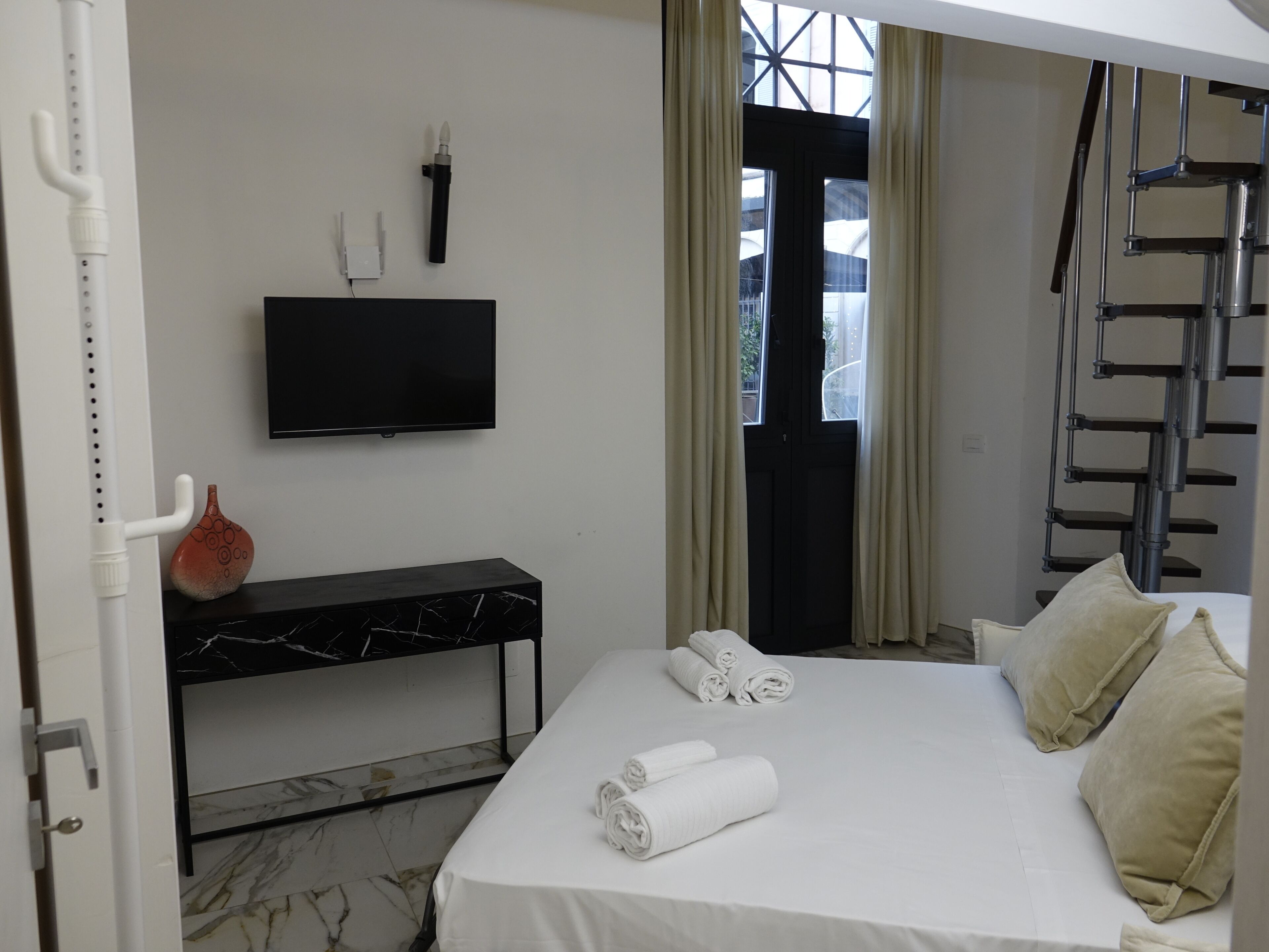 Habitación de hotel con cama, televisor de pantalla plana montado en la pared, una mesita consola con diseño de mármol y una ventana con cortinas.