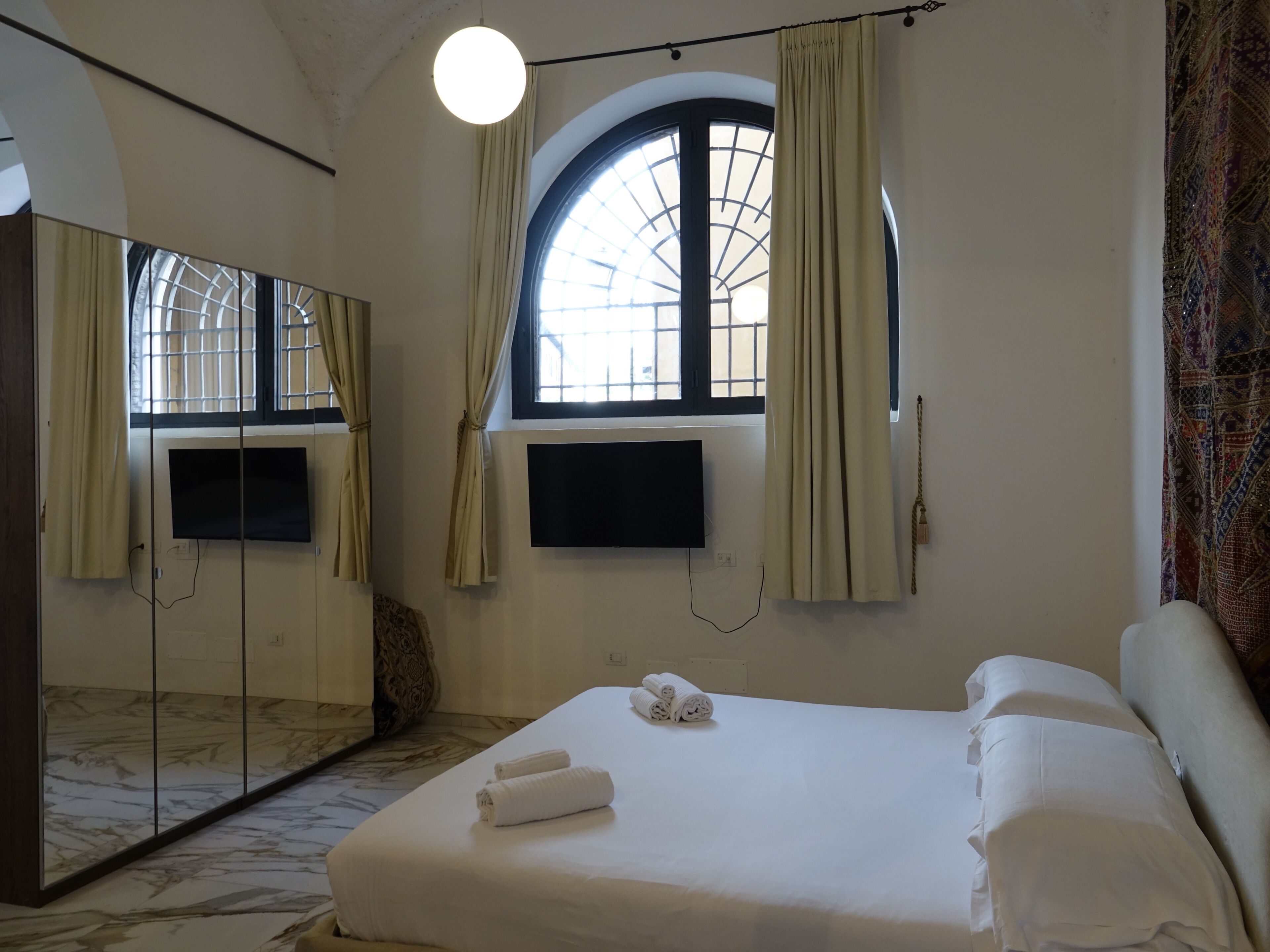 Een hotelkamer met een bed, een aan de muur bevestigde televisie, een grote spiegel en een raam met gordijnen.