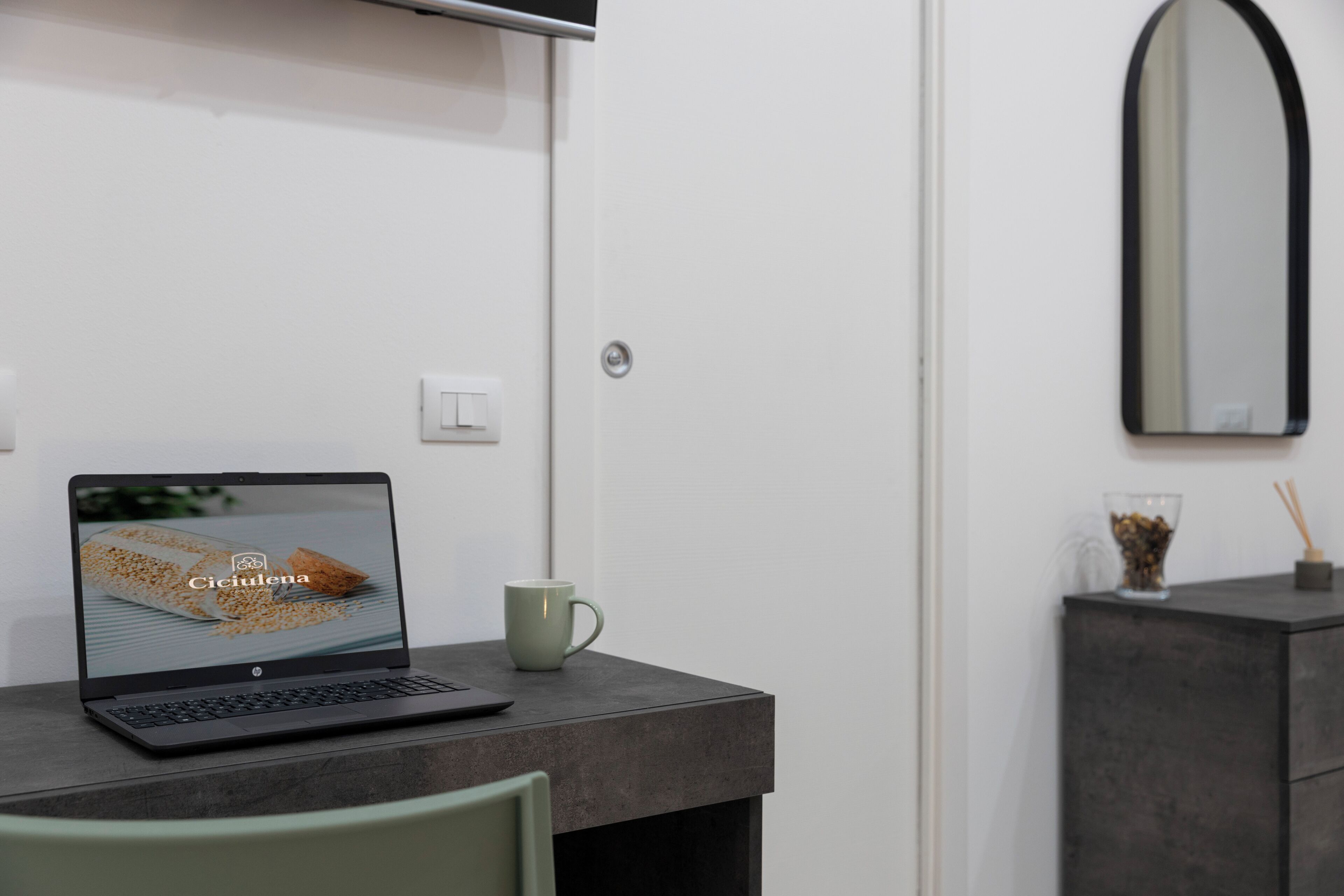 Ein Laptop auf einem Schreibtisch mit einer Tasse daneben, ein Spiegel an der Wand und eine kleine Kommode mit einem Glas und einer Kerze.