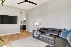 A modern living room with a leather sofa, a flat-screen TV mounted on the wall, a ceiling fan, and a kitchen area in the background. - Private Resort Backyard + Heated Pool & Spa + ¾-Acre Estate | Near The Boulders (Scottsdale)