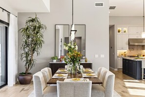 Un comedor moderno con una mesa de madera dispuesta para dos personas, rodeada de sillas tapizadas. Una alta planta en maceta se encuentra a la izquierda de la mesa.