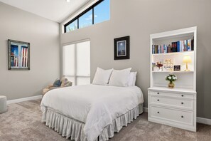 Ein Schlafzimmer mit einem großen Bett, einer weißen Kommode, einem Bücherregal und einem Fenster mit Blick auf den Himmel.