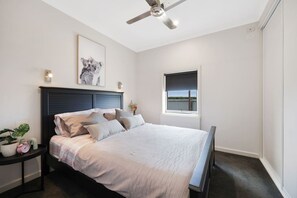 Una camera da letto con un letto, un comodino, una ventola a soffitto, una finestra e un quadro appeso al muro.