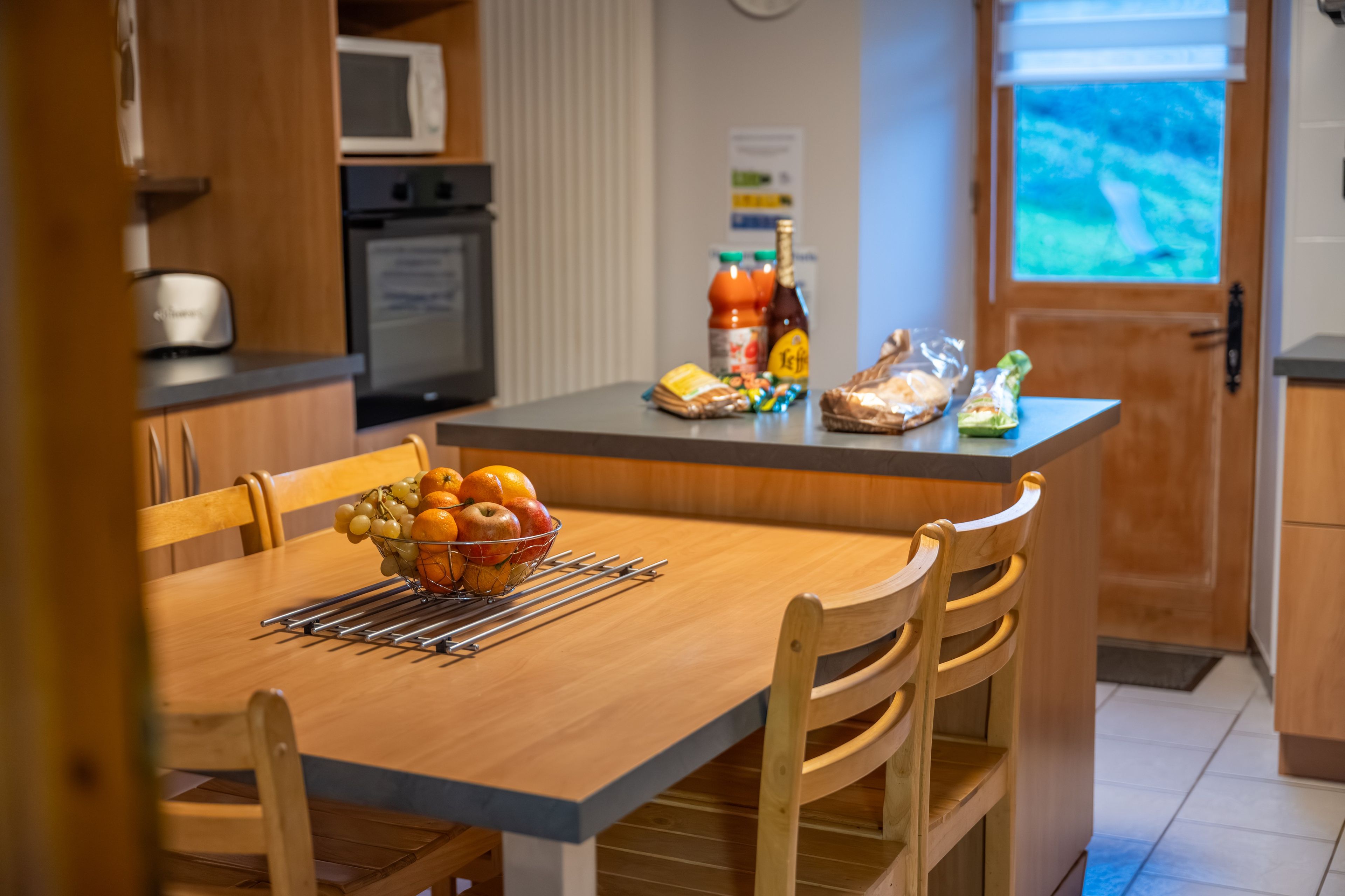 Une cuisine équipée d’une table à manger, de chaises, d’un panier de fruits, d’un four à micro-ondes, d’un réfrigérateur et d’une fenêtre donnant sur un paysage agréable.