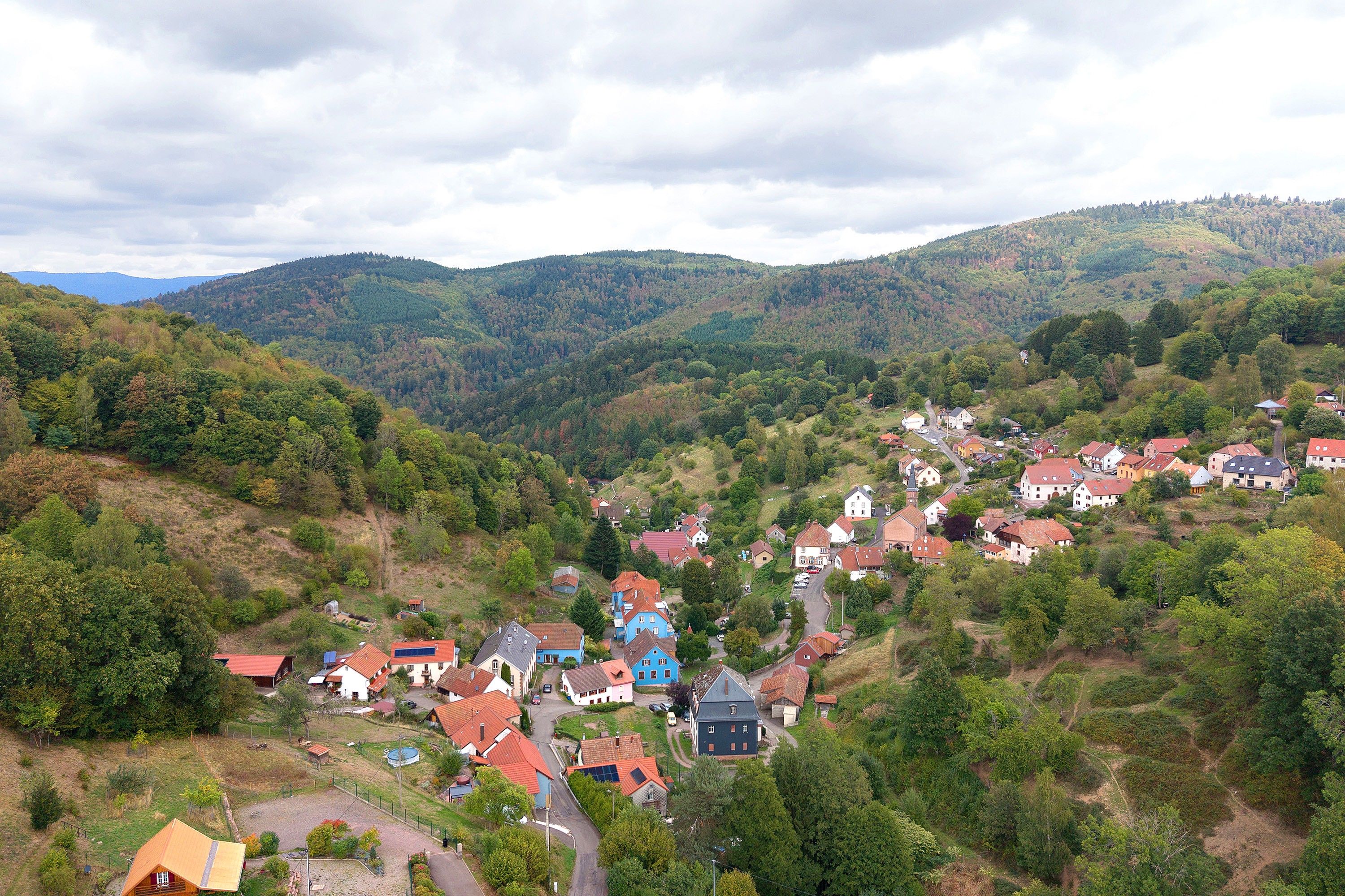 A hillside village with traditional houses, a winding road, and a backdrop of forested hills.
