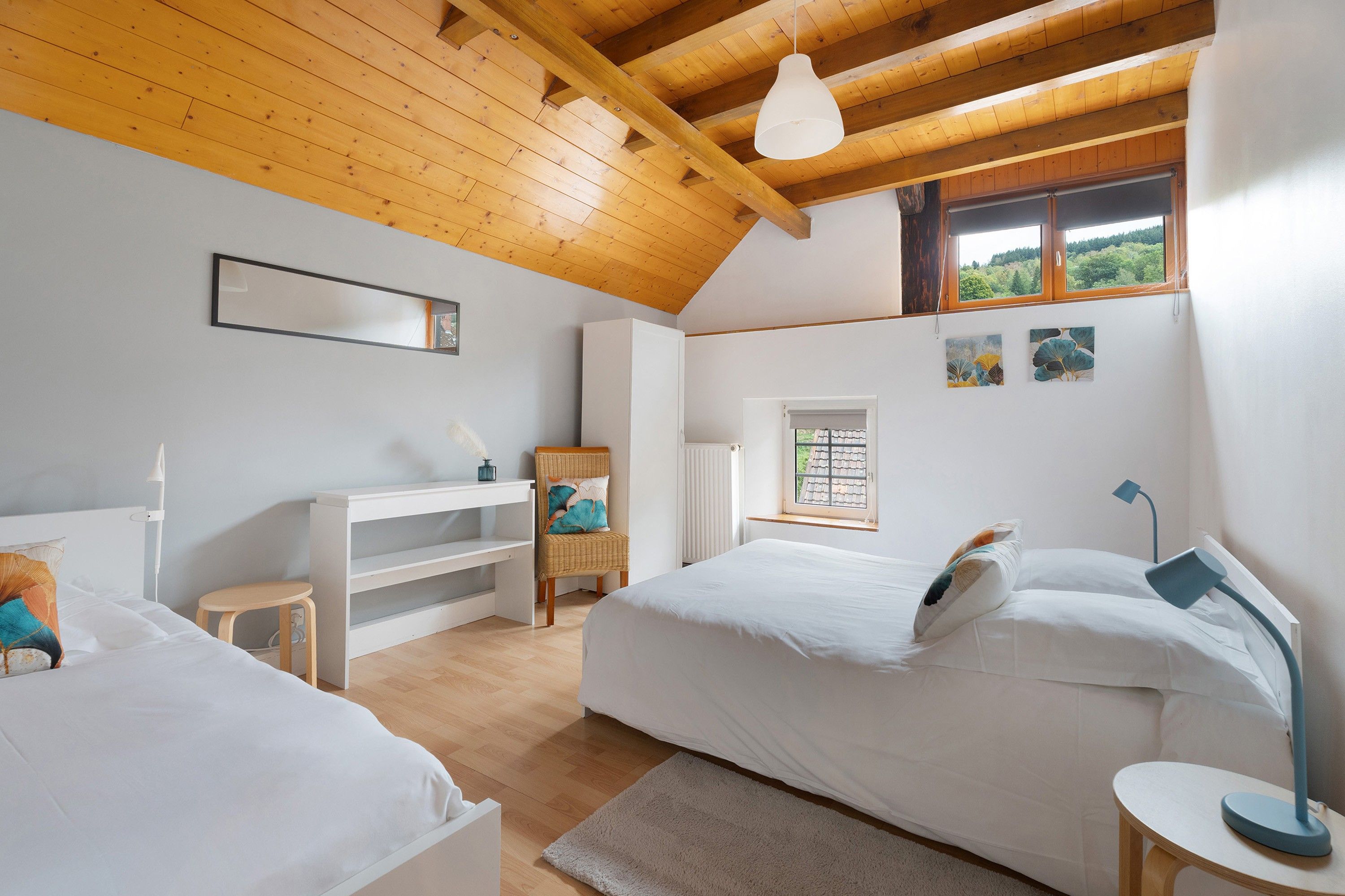 A bedroom with two beds, a desk, a chair, a radiator, and a window with a view of greenery.