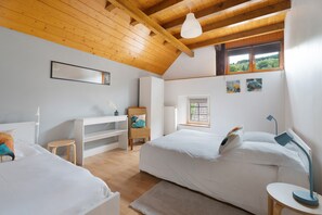 A bedroom with two beds, a desk, a chair, a radiator, and a window with a view of greenery.