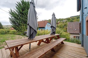 A wooden deck with picnic tables and umbrellas, surrounded by greenery and houses.