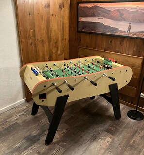 A foosball table in a room with wooden paneling and a television displaying a scenic landscape.