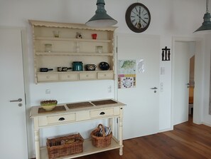 Ein Zimmer mit weißer Tür, einer Wanduhr, einem Regal mit diversen Gegenständen, einem Tisch mit Körben und einer Pendelleuchte.
