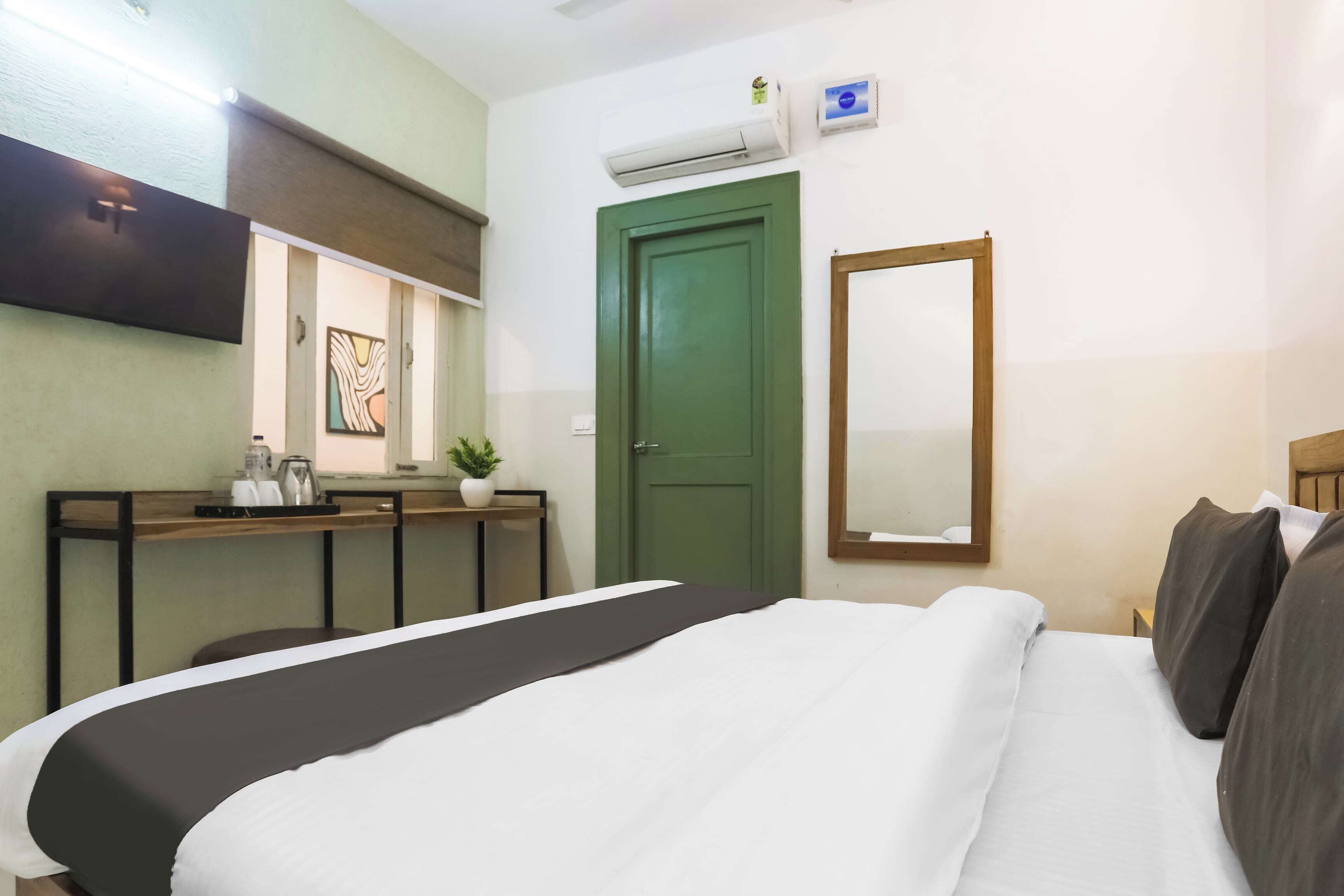 A hotel room with a bed, a desk with a computer, a green door, and an air conditioning unit.