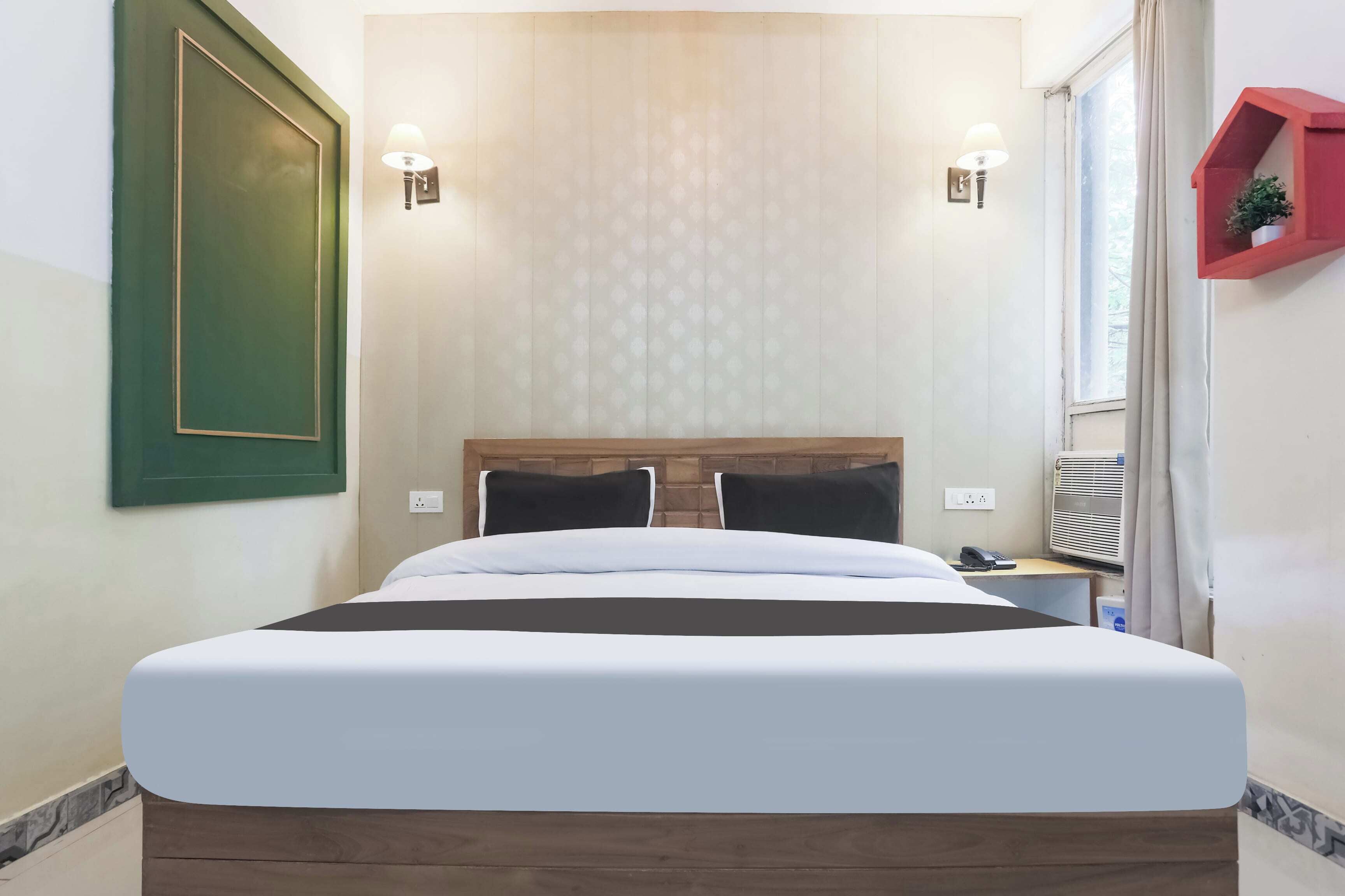 A hotel room with a large bed, two bedside lamps, a red plant shelf, and a window with curtains.