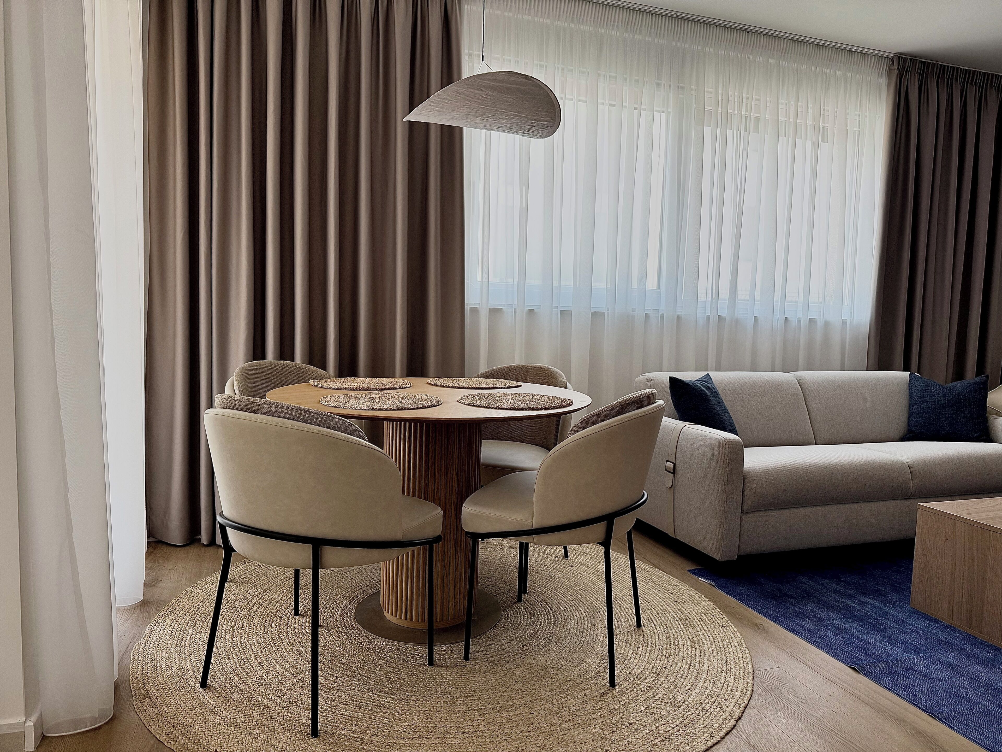 A modern living room with a round wooden table, beige sofas, and a blue rug.