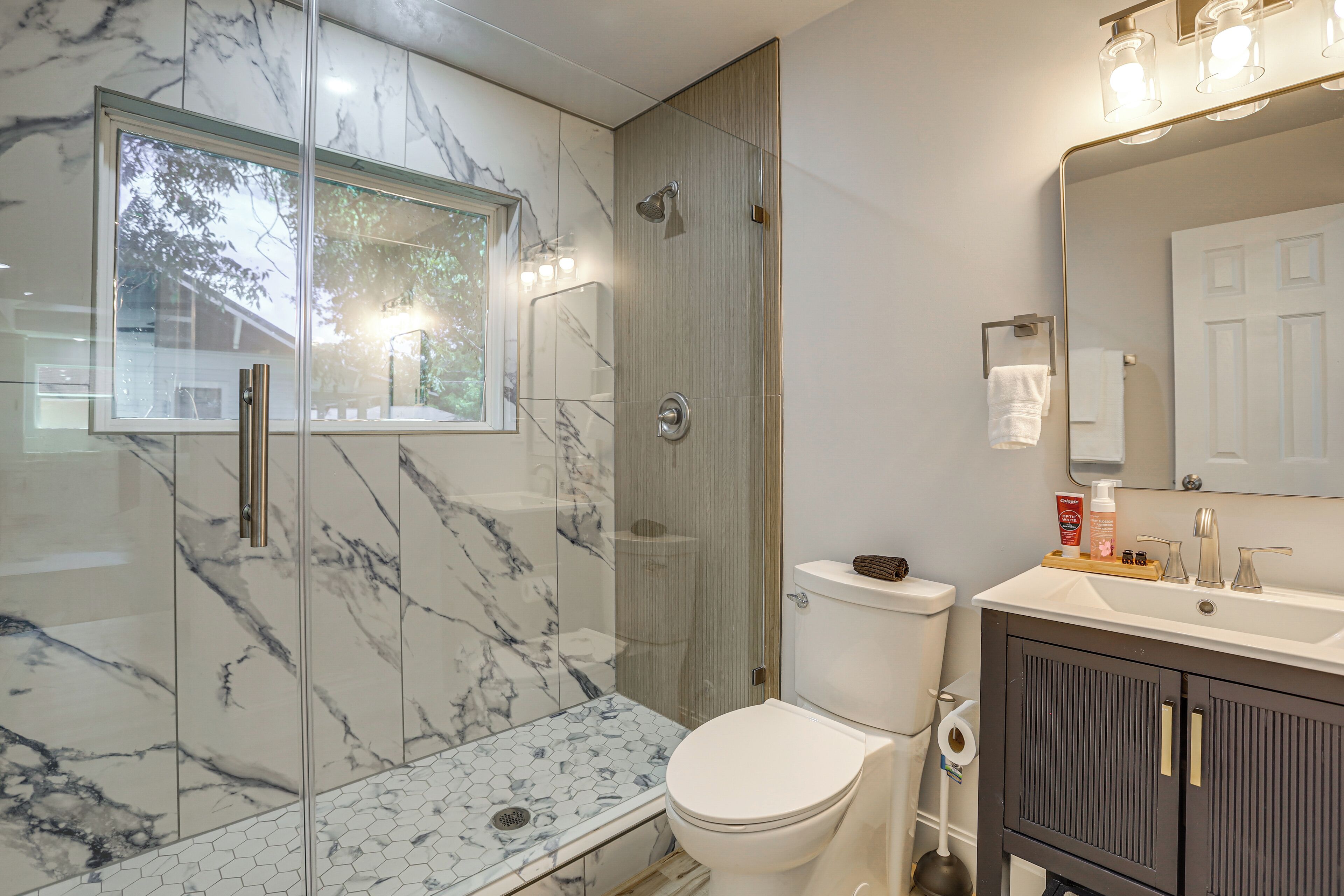 A bathroom with a glass-enclosed shower, a toilet, a sink with toiletries, and a mirror.