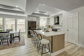 A modern kitchen with a central island, bar stools, and a dining area with a table and chairs. - Walk to Main St Tomball! Family Retreat w/ Yard (Tomball)