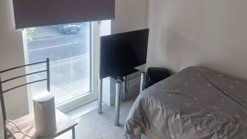 A bedroom with a bed, a chair, a desk, a television, and a window with a blind.