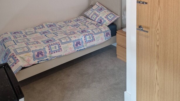 A single bed with a patterned bedspread, a small bedside table, a nightstand, a wooden door, and a wall-mounted shelf with potted plants. - A friendly budget room that delivers comfort stay (Woolston)