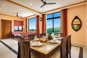 Un comedor con una mesa dispuesta para cuatro, ventanas amplias con vistas, un sofá y un reloj en la pared.