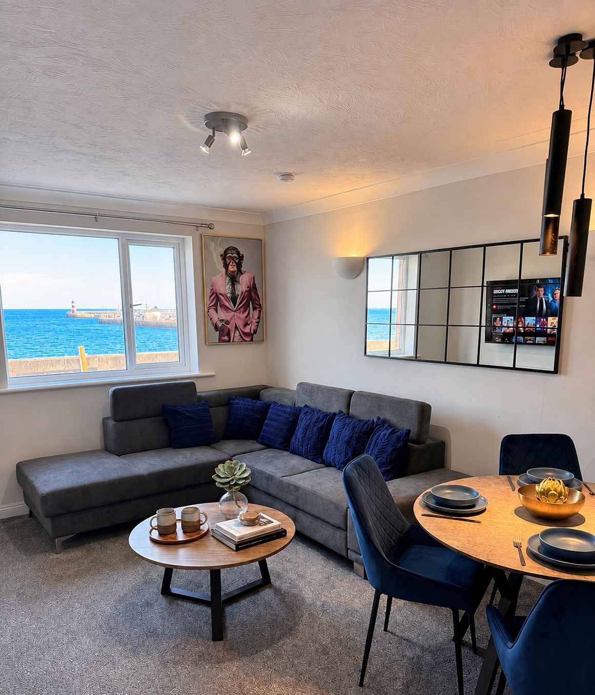 Ein modernes Wohnzimmer mit einem grauen Sofa, blauen Kissen, einem Couchtisch mit Büchern und einer Pflanze, sowie einem Essbereich mit einem runden Tisch und blauen Stühlen.