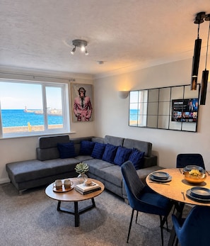 A modern living room with a grey sofa, blue cushions, a coffee table with books and a plant, and a dining area with a round table and blue chairs. - Stunning Sea View Flat on The Leas – Panoramic Estuary Sunsets, Steps to Beach (Minster on Sea)