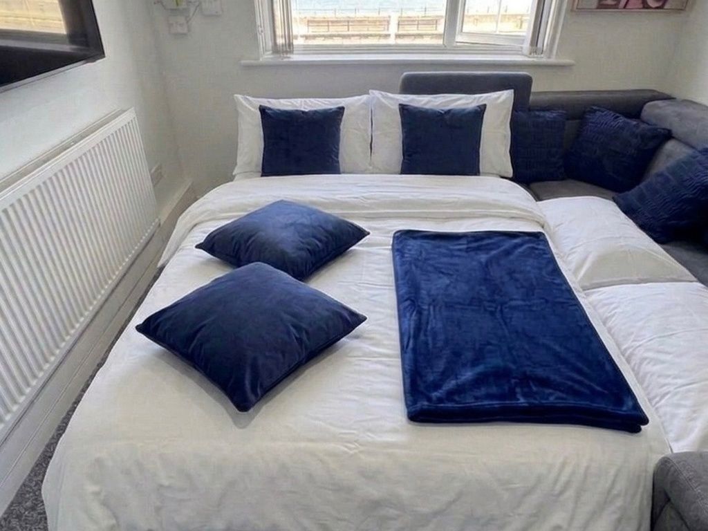 En seng med hvide sengetøj og blå puder, en grå sofa og et vindue med udsigt.