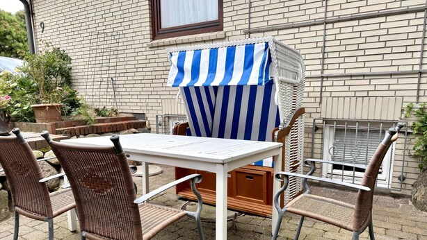 En uteplats med ett vitt bord och flätade stolar, en randig markis och en tegelvägg i bakgrunden.