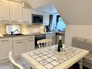 A compact kitchen with white cabinetry, a built-in microwave, and a dining area with a table featuring a patterned tablecloth. - Puan-Stöven-Weg, Apartment 1 - Cozy 4-room vacation apartment for 4 people, (Sylt)