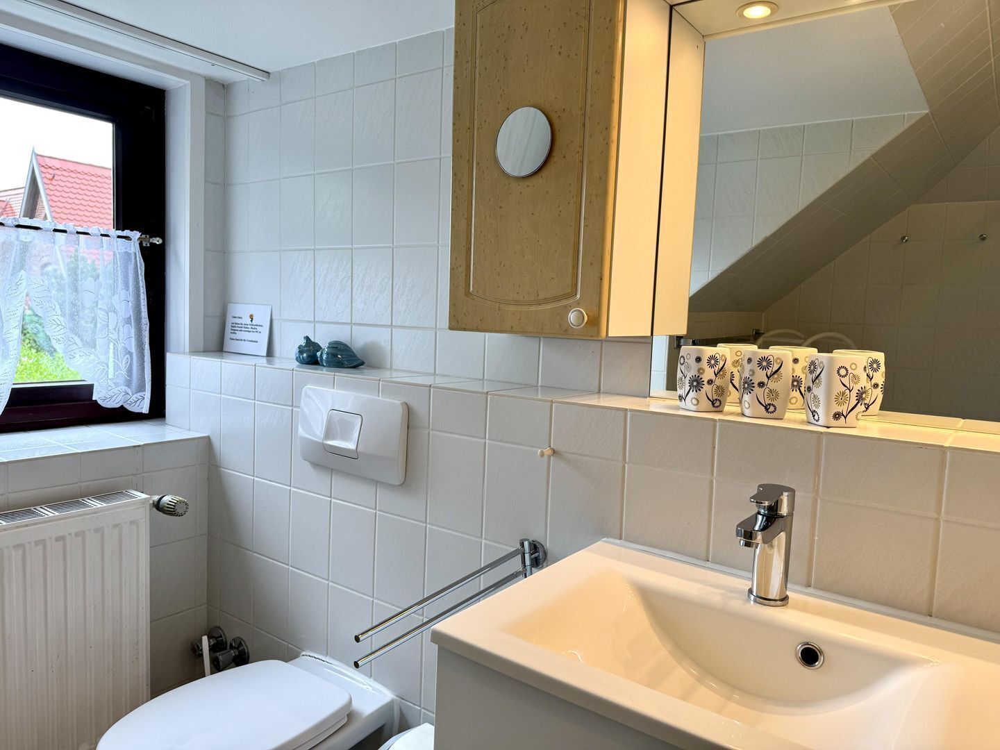 Une salle de bain avec un lavabo blanc, des toilettes, une fenêtre avec un rideau et un miroir au-dessus du lavabo.
