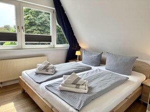 Ein ordentlich bezogenes Bett mit gestreifter Bettwäsche und Kissen, eine Nachttischlampe und ein Fenster mit Vorhängen.