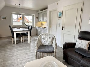 A dining area with a table and chairs, a wicker chair with a cushion, a brown leather sofa with a cushion, and a kitchen in the background. - Kampstraße, Apartment 3a - Cozy 3-room vacation apartment with a balcony in a quiet (Sylt)