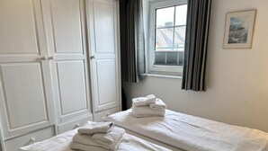 A room with two beds, each with white linens and towels, a window with curtains, and a framed picture on the wall. - Kampstraße, Apartment 3a - Cozy 3-room vacation apartment with a balcony in a quiet (Sylt)