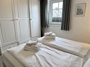 A room with two beds, each with white linens and towels, a window with curtains, and a framed picture on the wall. - Kampstraße, Apartment 3a - Cozy 3-room vacation apartment with a balcony in a quiet (Sylt)