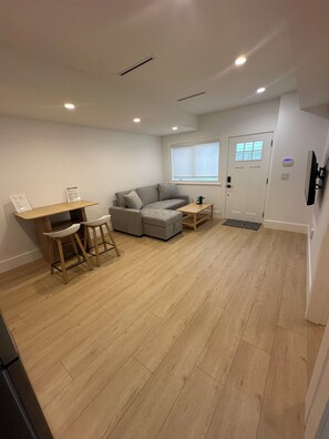 A modern living room with a grey sectional sofa, a wooden dining table with two chairs, a small wooden coffee table, and a white door with a window. - Cozy Burnaby Heights Suite Private 1BR + 1 Bath (Burnaby)